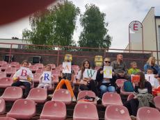 Grupa dzieci siedzi na stadionie każde dziecko w ręku trzyma kartkę z jedną literą z czego powstaje kolorowany napis brawo.jpg