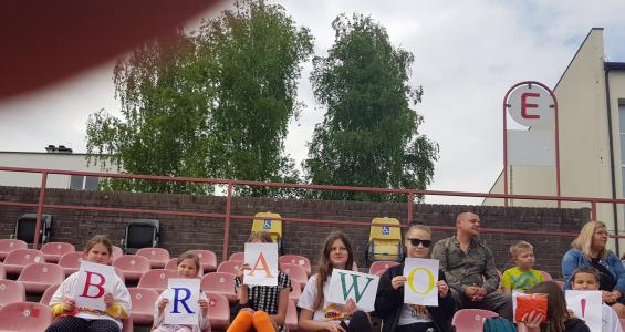 Grupa dzieci siedzi na stadionie każde dziecko w ręku trzyma kartkę z jedną literą z czego powstaje kolorowany napis brawo.jpg