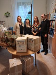 Na pierwszym planie widać trzy kartonowe paczki ustawione na drewnianym stoliku. Za paczkami w gabinecie Dyrektora Domu Dziecka w Cieszynie stoją uśmiechnięci od lewej strony 3 kobiety i jeden mężczyzna - są to od lewej dyrektorka Domu Dziecka w Cieszynie oraz trójka darczyńców. 