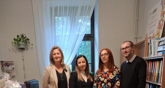 Na pierwszym planie widać trzy kartonowe paczki ustawione na drewnianym stoliku. Za paczkami w gabinecie Dyrektora Domu Dziecka w Cieszynie stoją uśmiechnięci od lewej strony 3 kobiety i jeden mężczyzna - są to od lewej dyrektorka Domu Dziecka w Cieszynie oraz trójka darczyńców. 